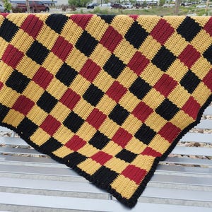 May include: A handmade crochet blanket with a checkerboard pattern in black, gold, and burgundy. The blanket is folded over a bench, showcasing the detailed stitch work and color arrangement. The edges are finished with a black border.