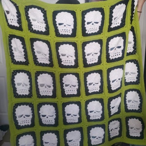 May include: A green and black crocheted blanket with a pattern of white skulls. The blanket is made of yarn and has a soft, fuzzy texture.