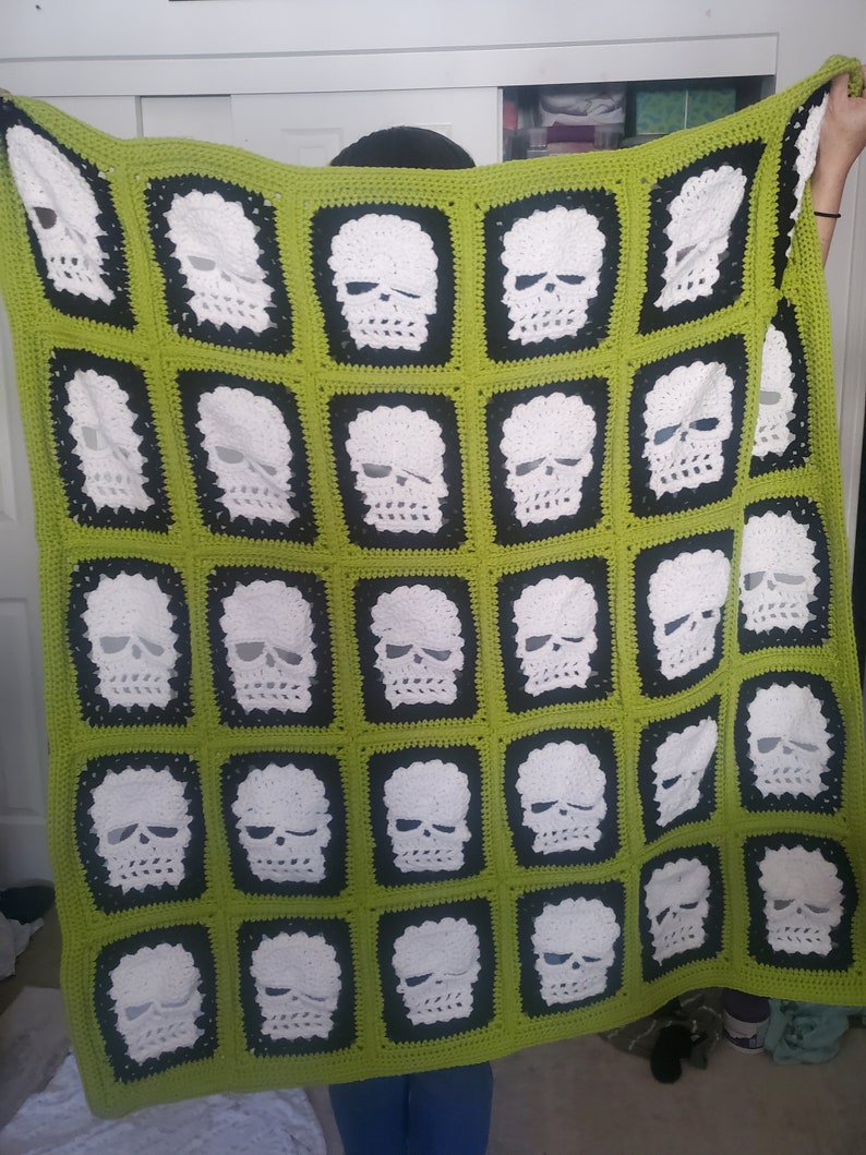 May include: A green and black crocheted blanket with a pattern of white skulls. The blanket is made of yarn and has a soft, fuzzy texture.