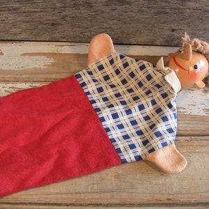 May include: A hand puppet with a wooden head, brown hair, and a blue and white plaid shirt. The puppet is wearing a red and white plaid skirt and has a red felt hand.