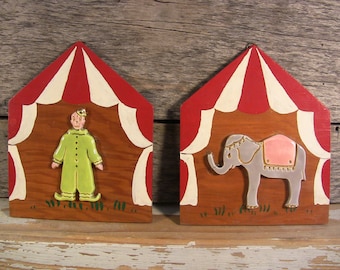 May include: Two wooden circus tent plaques with painted details. One plaque features a clown with a green suit and a red hat. The other plaque features a gray elephant with a pink blanket.