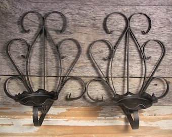 May include: Two wrought iron wall sconces. Each sconce features a decorative, symmetrical design with a central vertical element and curved accents. The sconces have a dark, possibly antique, finish and a circular candle holder.