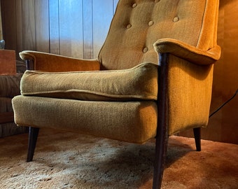 May include: A mustard yellow armchair with a tufted back and dark wood legs. The chair is sitting on a beige shag carpet.