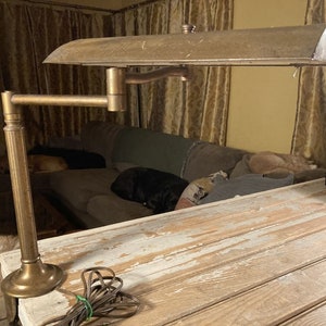 May include: An antique brass desk lamp with an adjustable arm and a curved shade. The lamp is mounted on a wooden desk with a distressed finish. The lamp's cord is visible, and the background includes a sofa and curtains.