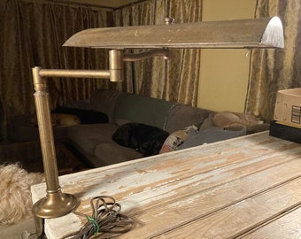 May include: An antique brass desk lamp with an adjustable arm and a curved shade. The lamp is mounted on a wooden desk with a distressed finish. The lamp's cord is visible, and the background includes a sofa and curtains.