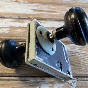 May include: An antique door knob set with a black finish. The set includes two round knobs and a rectangular backplate with a keyhole. The metal hardware has a distressed appearance, suggesting age and use.
