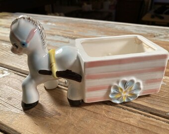 May include: A white ceramic planter shaped like a horse pulling a wagon. The wagon has pink and white stripes and a blue flower on the wheel.