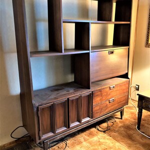 May include: A large, dark brown wooden bookcase with a built-in desk and drawers. The bookcase has multiple shelves and compartments, and the desk has a drop-down door. The bookcase is standing on a brown carpet.