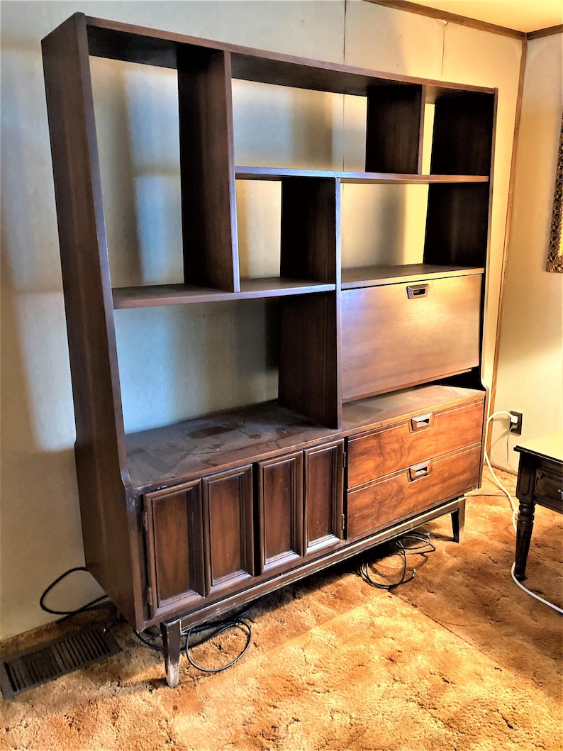 May include: A large, dark brown wooden bookcase with a built-in desk and drawers. The bookcase has multiple shelves and compartments, and the desk has a drop-down door. The bookcase is standing on a brown carpet.