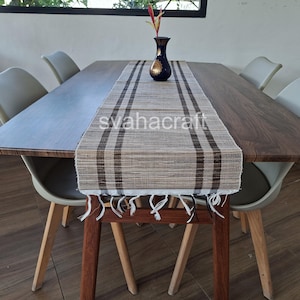 May include: A rectangular table runner with a brown and beige plaid pattern, featuring fringed edges on the ends. The runner is placed on a dark wood dining table with four light gray chairs. A dark blue vase with gold accents sits on top.