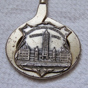 May include: A silver-toned souvenir spoon featuring a detailed relief of Duluth Central High School in Duluth, Minnesota. The spoon's bowl is shaped like a shield with the school's name and location engraved.