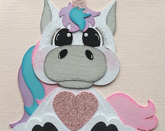 May include: A whimsical papercraft unicorn with a glittery horn and heart. The unicorn features a white body with black hooves, a gray face, and a mane in shades of pink, blue, and purple. The design is layered and textured.