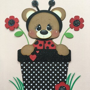 May include: A brown teddy bear dressed as a ladybug is sitting in a black and white polka dot flower pot. The pot has green grass and red flowers around it.