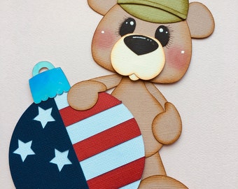 Op de afbeelding: Een bruine teddybeer met een groene hoed houdt een rood, wit en blauwe kerstbal vast met sterren. De kerstbal is aan de ene kant versierd met een rood, wit en blauw gestreept patroon en aan de andere kant met een blauwe achtergrond met witte sterren.