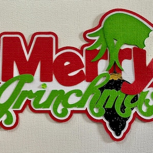 May include: A layered papercraft design with the words "Merry Grinchmas" in red and green. The design includes a green hand and a black ornament, all outlined in white.