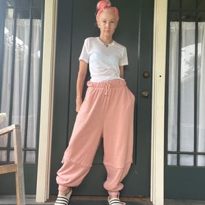 May include: A person wearing a white t-shirt and light pink sweatpants with a drawstring waist and large pockets. The person is also wearing striped sandals. The background is a dark green door and white curtains.