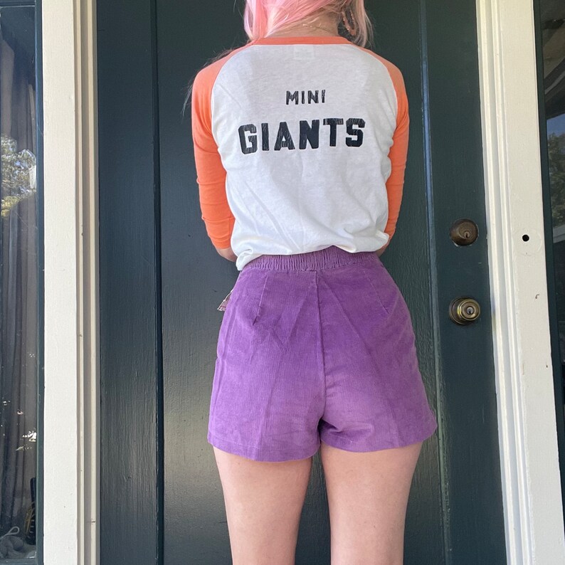 May include: A white and orange baseball jersey with the text "MINI GIANTS" on the back. The person is wearing purple corduroy shorts.