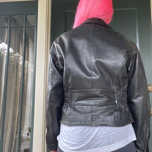 May include: Black leather jacket with a cinched waist and a zipper closure. The jacket has a cropped length and a classic biker style.