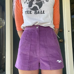 May include: Purple corduroy shorts with a white embroidered bird on the right leg. The shorts have a high waist and a button closure.