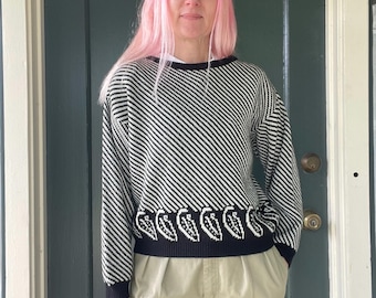 May include: Black and white patterned sweater with a crew neck and long sleeves. The sweater features a diagonal stripe design and a leaf pattern along the bottom hem. The person is wearing khaki pants.