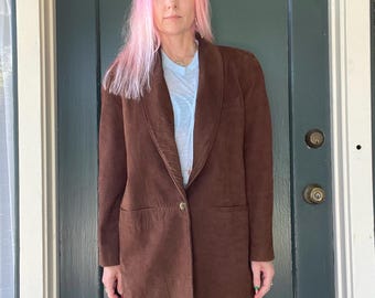 May include: A brown suede blazer with a notched lapel and two front pockets. The blazer has a single-button closure and a relaxed fit. The model is wearing a light blue t-shirt underneath the blazer.
