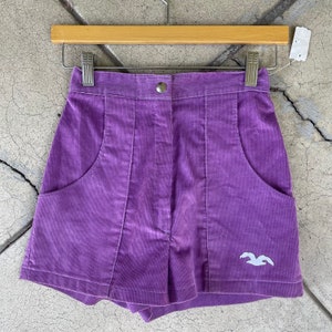 May include: Purple corduroy shorts with a white bird logo on the right leg. The shorts have two front pockets and a button closure.