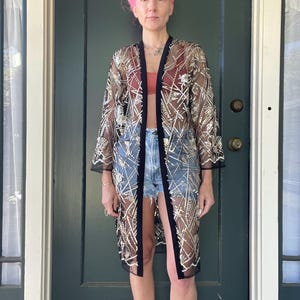 May include: A sheer black kimono-style jacket with gold and white geometric embroidery. The jacket has a black trim and is worn open. The garment is knee-length and has long sleeves. The model is wearing denim shorts and a red top.