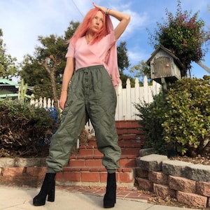 May include: A woman wearing a pink wrap top and green quilted pants. She is standing on a set of brick steps with a white picket fence behind her.