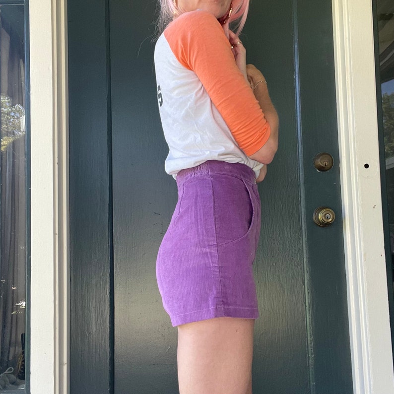 May include: A woman wearing a white and orange baseball tee shirt with a purple corduroy short. The shirt has the word "6" printed on the back.