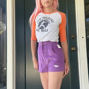 May include: A woman wearing a white and orange baseball tee shirt with the text "San Marino Tee Ball" and a purple corduroy short with a white bird logo.
