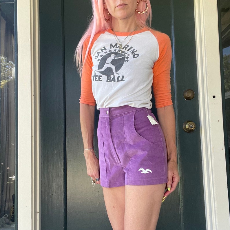 May include: A woman wearing a white and orange baseball tee shirt with the text "San Marino Tee Ball" and a purple corduroy short with a white bird logo.