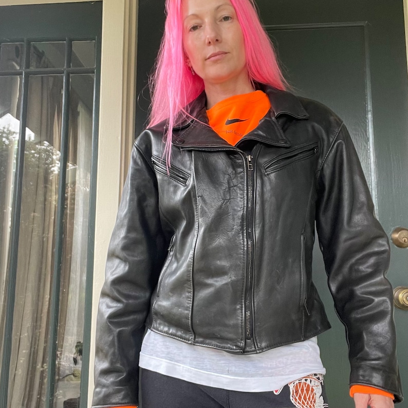 May include: A black leather motorcycle jacket with a zipper closure and a stand-up collar. The jacket has a white t-shirt underneath and a bright orange shirt with a black Nike swoosh logo.