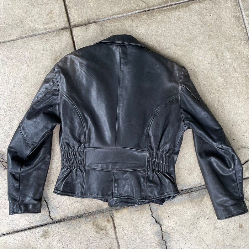 May include: Black leather jacket with a cinched waist and a zipper closure. The jacket has a collar and long sleeves.