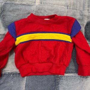 May include: A red long-sleeved sweatshirt with a blue and yellow stripe design across the chest. The shoulders are blue. The sweatshirt is made of a soft material and has a ribbed collar and cuffs. The garment is likely for a child.