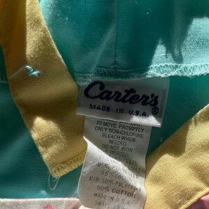 May include: A close-up of a baby's bib with a tag that reads "Carter's MADE IN U.S.A." The bib is a mix of colors, including light blue, yellow, pink, and white with pink polka dots. The tag provides care instructions and fabric content.