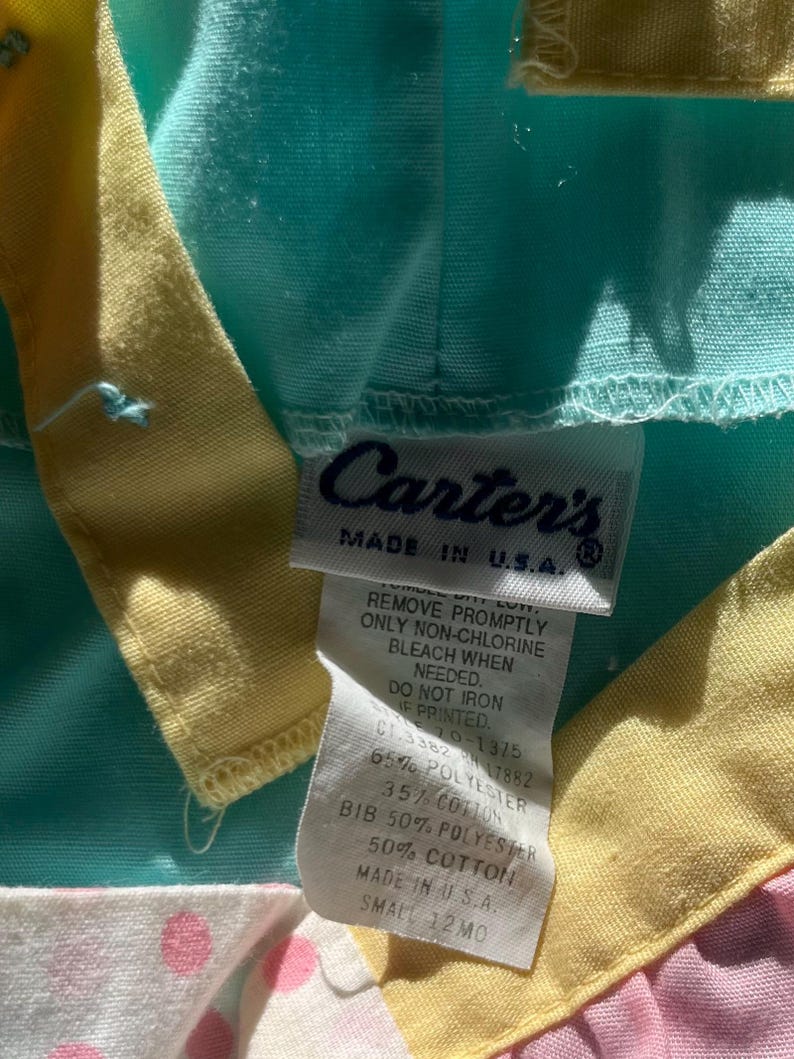 May include: A close-up of a baby's bib with a tag that reads "Carter's MADE IN U.S.A." The bib is a mix of colors, including light blue, yellow, pink, and white with pink polka dots. The tag provides care instructions and fabric content.
