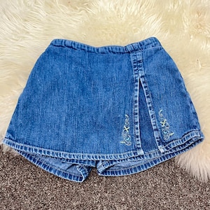 May include: Blue denim skort with a front slit and embroidered floral details. The skort features a denim skirt overlay with attached shorts underneath. The embroidery includes light green and blue thread. The skort is designed for children.
