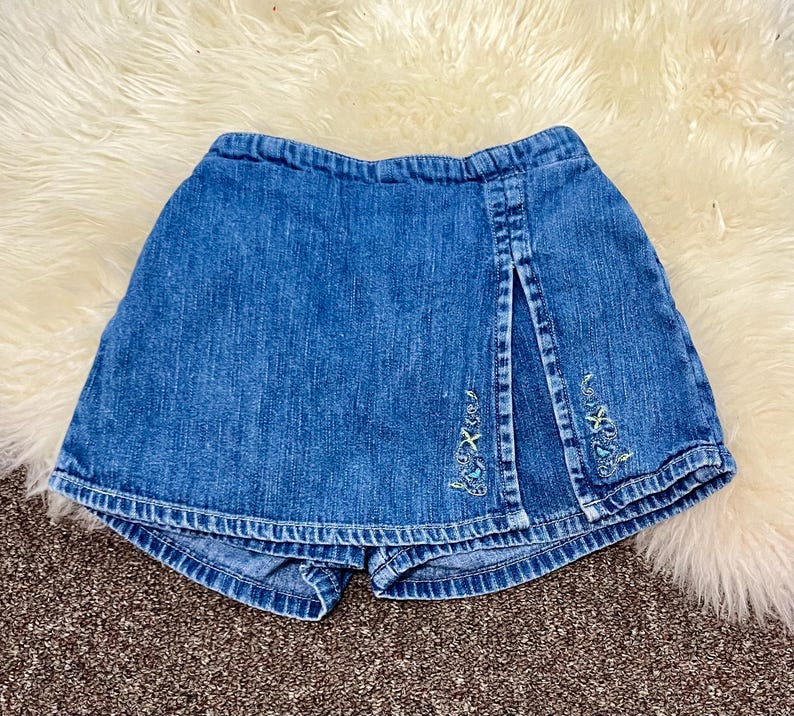 May include: Blue denim skort with a front slit and embroidered floral details. The skort features a denim skirt overlay with attached shorts underneath. The embroidery includes light green and blue thread. The skort is designed for children.