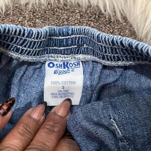 May include: Close-up of a blue denim garment with an elastic waistband. A white tag displays the brand "OshKosh B'gosh" and details: "100% Cotton", size "5", and "Made in Philippines". The garment's texture is visible.