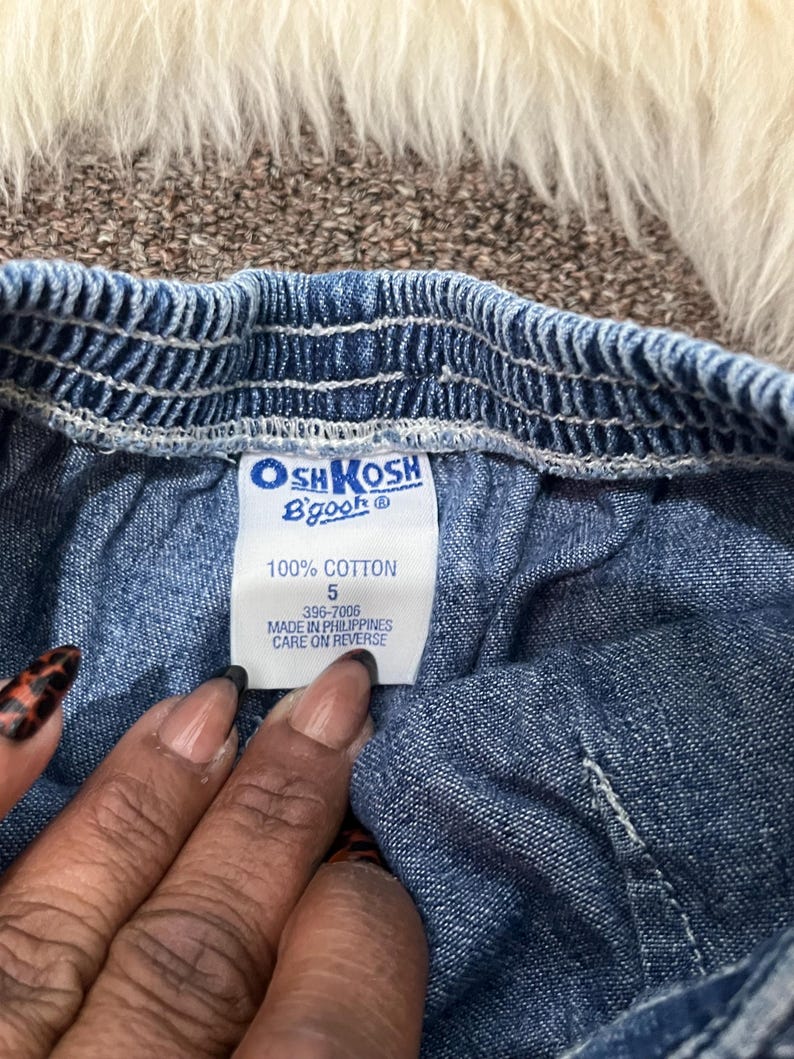 May include: Close-up of a blue denim garment with an elastic waistband. A white tag displays the brand "OshKosh B'gosh" and details: "100% Cotton", size "5", and "Made in Philippines". The garment's texture is visible.