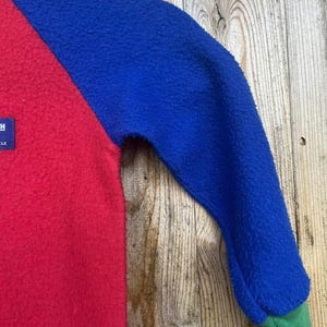 Puede incluir: Un jersey de forro polar con bloques de color con cuerpo rojo, mangas azules y pu&ntilde;os verdes. La prenda tiene un aspecto texturizado y una peque&ntilde;a etiqueta en la parte roja. El fondo es una superficie de madera.