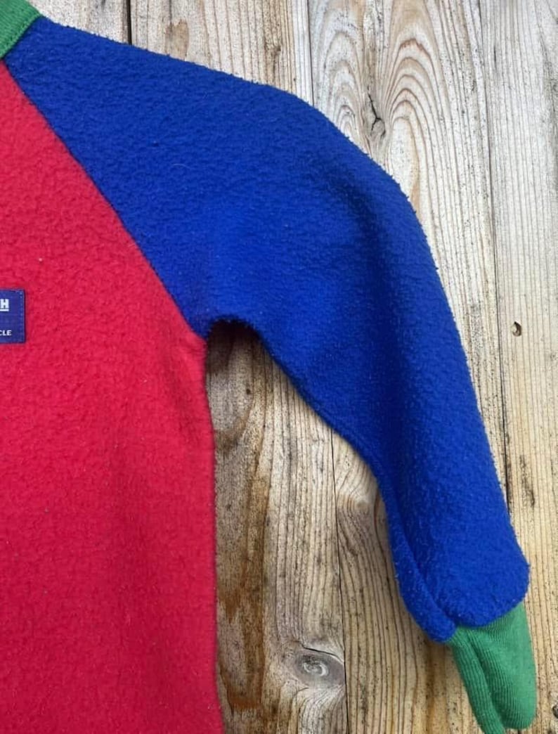 Puede incluir: Un jersey de forro polar con bloques de color con cuerpo rojo, mangas azules y pu&ntilde;os verdes. La prenda tiene un aspecto texturizado y una peque&ntilde;a etiqueta en la parte roja. El fondo es una superficie de madera.