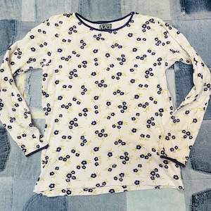 Peut inclure: T-shirt blanc à manches longues orné d'un motif floral. Le motif répète de petites fleurs bleues avec des feuilles vertes. L'encolure et les poignets sont bordés de bleu marine. L'étiquette indique "PLACE".