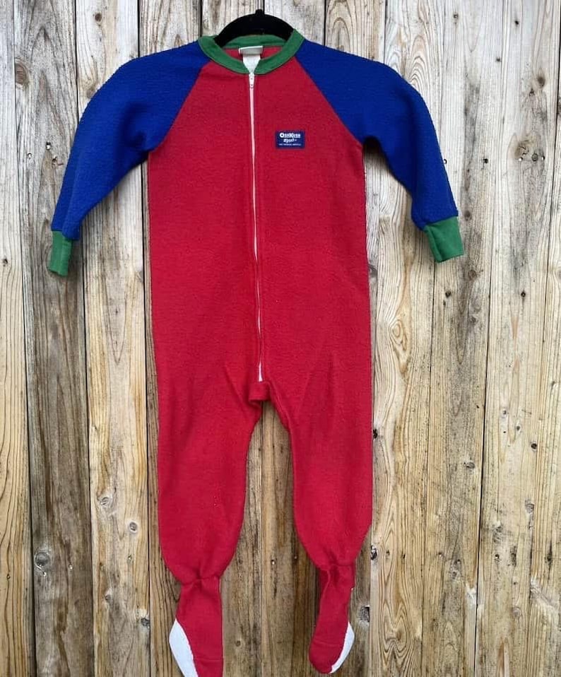 Puede incluir: Un pijama de una pieza rojo, azul y verde con cremallera completa. Las mangas y la parte superior del cuerpo son azules, con ribetes verdes alrededor del cuello y los pu&ntilde;os. Los pies son rojos con suelas blancas.