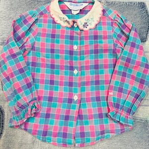 Puede incluir: Camisa vintage a cuadros en rosa, turquesa, morado y gris. Tiene mangas largas con puños con volantes y un cuello blanco con bordado floral. La camisa se abrocha con botones blancos. La marca "OshKosh" es visible.