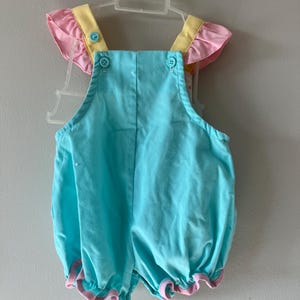 May include: A baby's romper in a light blue color, with pink and yellow accents. The romper has ruffled sleeves and leg openings, and is fastened with buttons. The garment is hanging on a white plastic hanger.