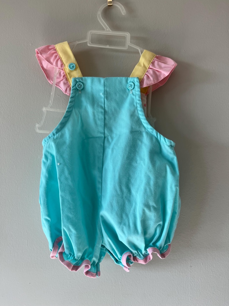 May include: A baby's romper in a light blue color, with pink and yellow accents. The romper has ruffled sleeves and leg openings, and is fastened with buttons. The garment is hanging on a white plastic hanger.