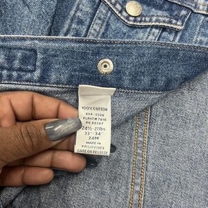 May include: Close-up of a denim garment with a label held by a hand. The label reads "100% COTTON" and includes measurements of 24 1/2 - 27 lbs and 33"-34". The garment has metal buttons and visible stitching.
