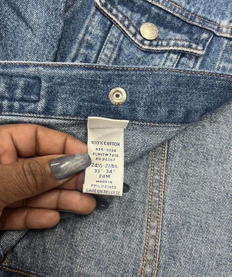 May include: Close-up of a denim garment with a label held by a hand. The label reads "100% COTTON" and includes measurements of 24 1/2 - 27 lbs and 33"-34". The garment has metal buttons and visible stitching.