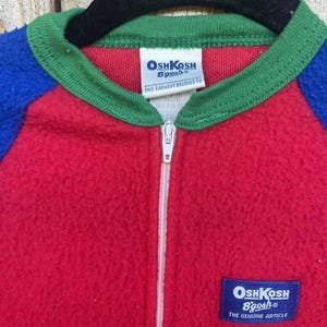 Puede incluir: Un mono vintage OshKosh B'gosh de una pieza en rojo, azul y verde. El mono tiene una cremallera completa, un cuello verde y mangas azules. La etiqueta OshKosh B'gosh es visible en el pecho. La prenda est&aacute; colgada en una percha negra.